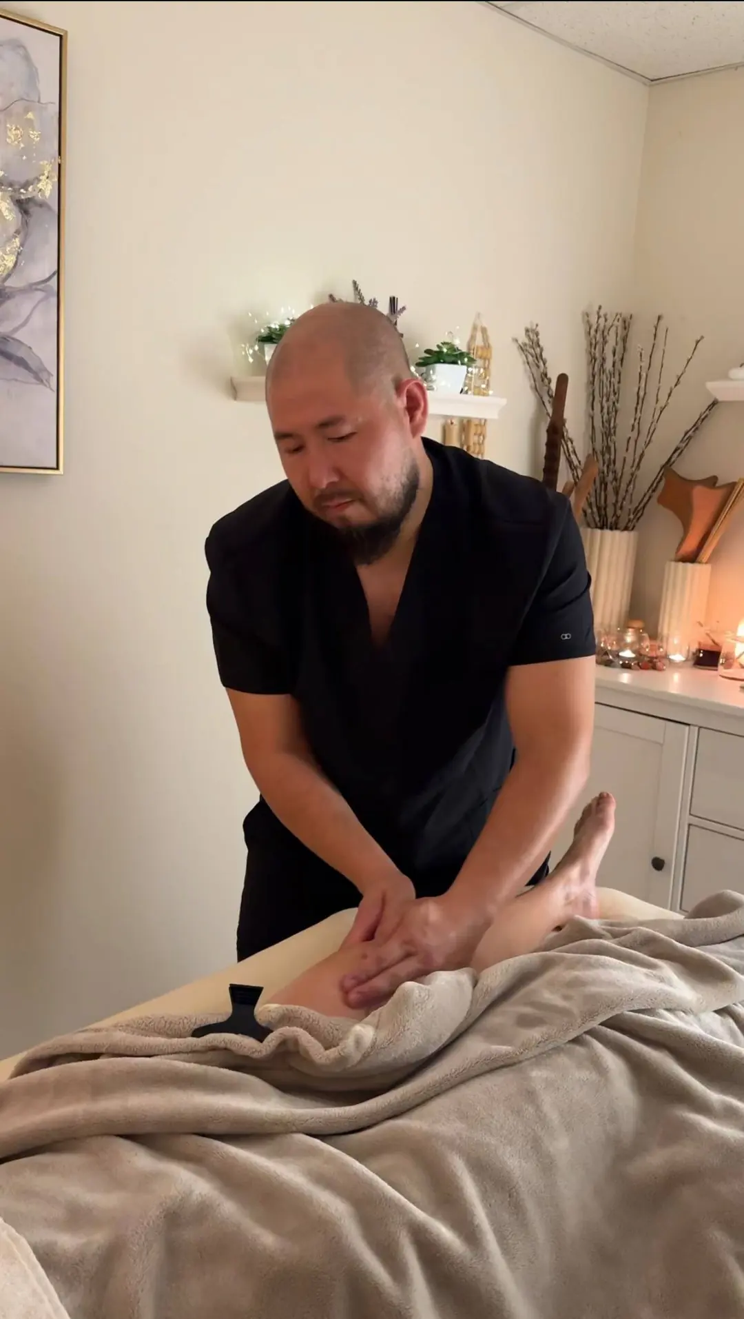 Tony in black scrubs massaging a client's leg under a soft blanket in a warmly lit room